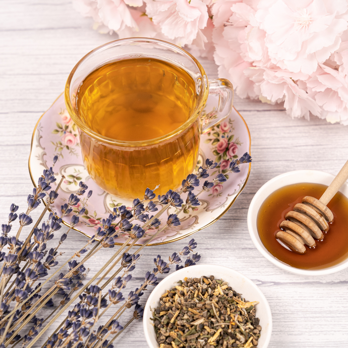 Cup of brewed Organic Menopause Tea in a floral teacup surrounded by lavender sprigs, honey, and loose herbal tea blend on a light wooden surface with pink flowers in the background.