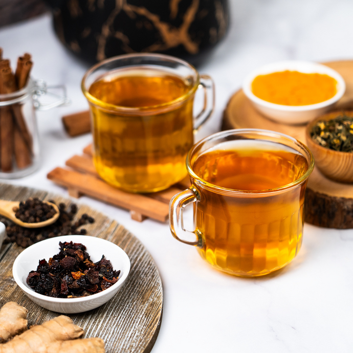 Two glass cups of golden anti-inflammatory tea surrounded by turmeric, cinnamon sticks, black pepper, dried berries, and ginger on a white and wooden surface.