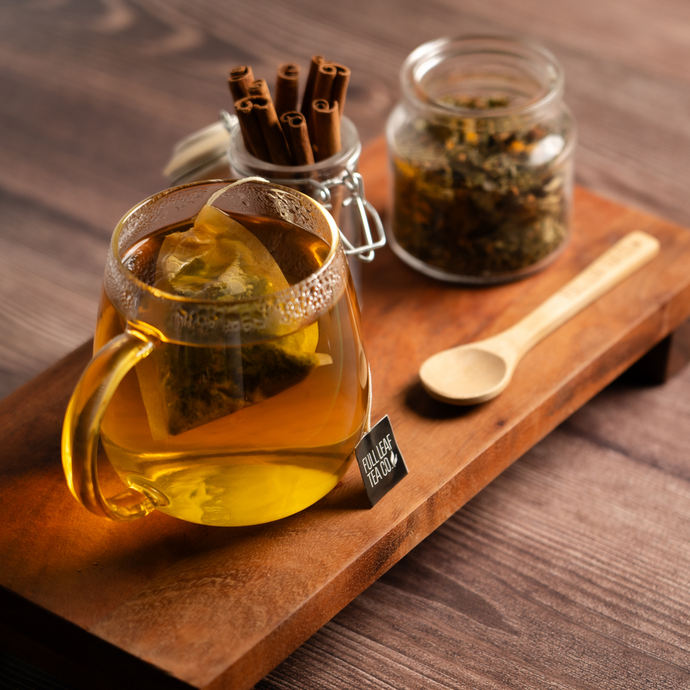 Glass mug of Organic Healthy Heart Tea with a tea bag steeping, placed on a wooden tray beside a jar of loose tea, cinnamon sticks, and a wooden spoon.