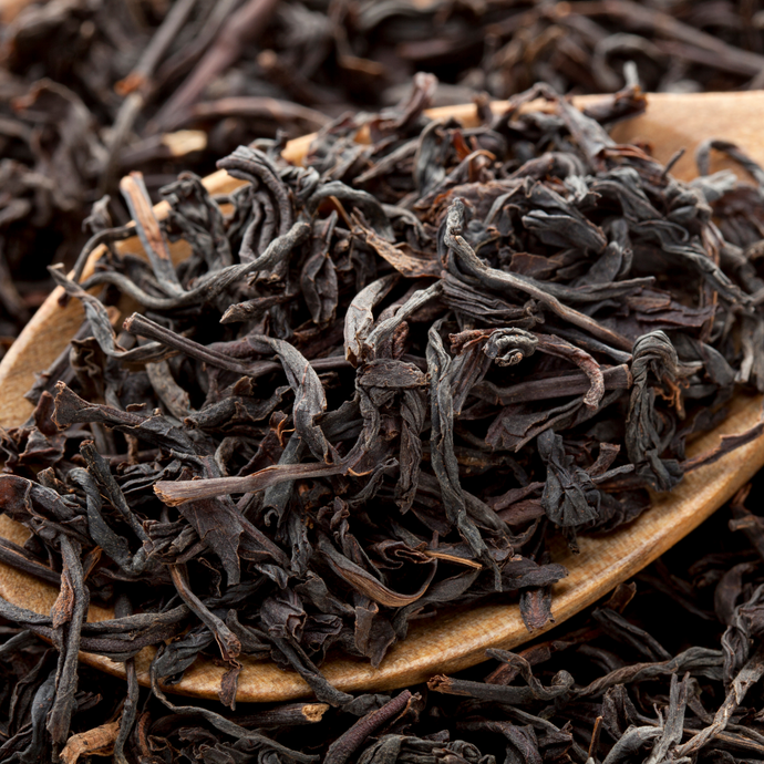 Close-up of loose Organic Assam black tea leaves in a wooden spoon, showing their rich dark brown color and twisted texture.