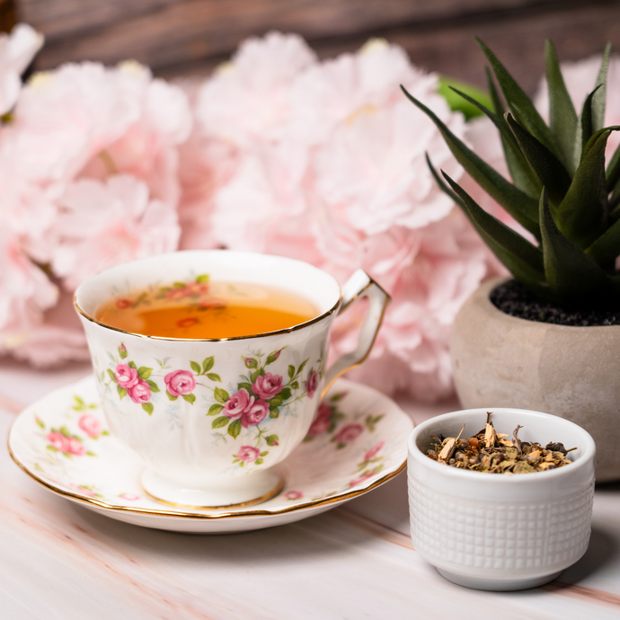 Cup of Organic Cycle Support Tea in a floral china teacup and saucer, with a small bowl of loose herbal blend beside it, set against soft pink flowers and a potted succulent on a light marble surface.
