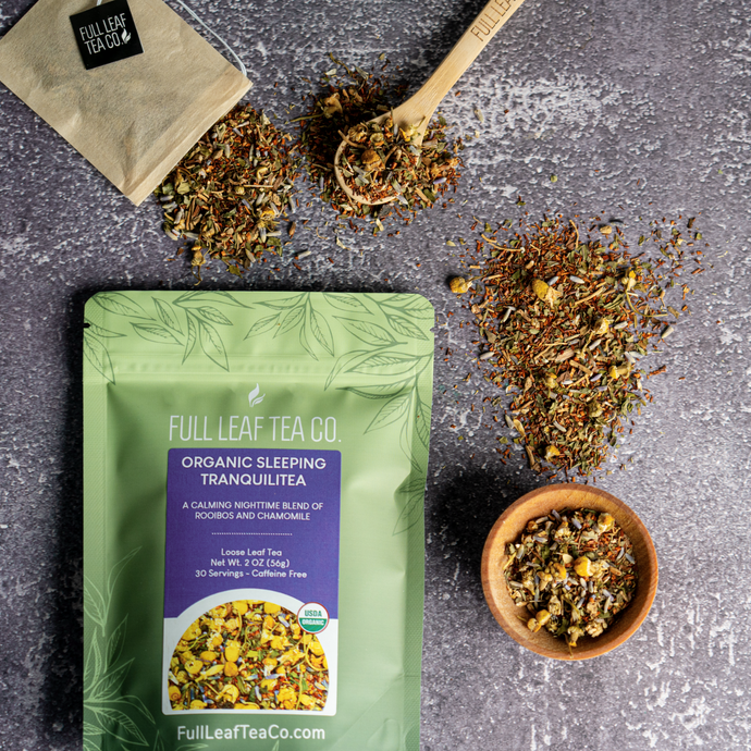 Full Leaf Tea Co. Organic Sleeping TranquiliTea package surrounded by loose leaf rooibos and chamomile blend, a bamboo tea spoon, and tea bag on a textured gray surface.