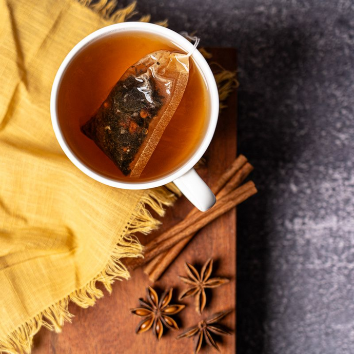 Cup of spiced tea with a tea bag steeping, placed on a wooden board with cinnamon sticks and star anise beside a yellow cloth on a dark stone background.