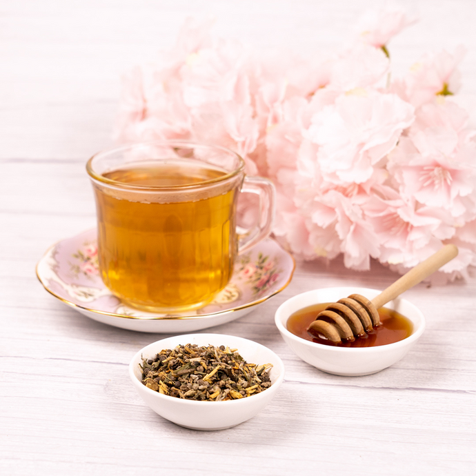 Glass teacup of Organic Menopause Tea on a floral saucer with bowls of loose herbal blend and honey, styled with soft pink flowers for a soothing and elegant presentation.