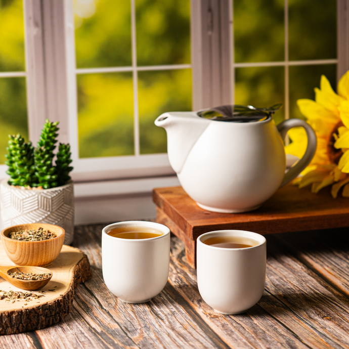 Two cups of Organic Dandelion Tea with a white teapot on a wooden table near a bright window, surrounded by a small succulent, wooden bowls of dried herbs, and yellow sunflowers in warm natural light.