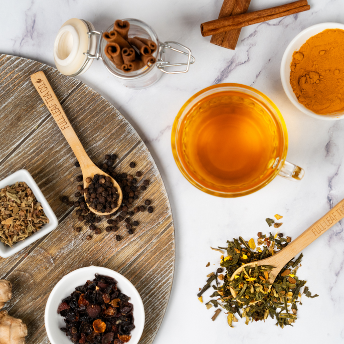 Cup of golden anti-inflammatory tea surrounded by loose leaf blend, turmeric powder, cinnamon sticks, black pepper, and dried herbs on a marble and wood surface.