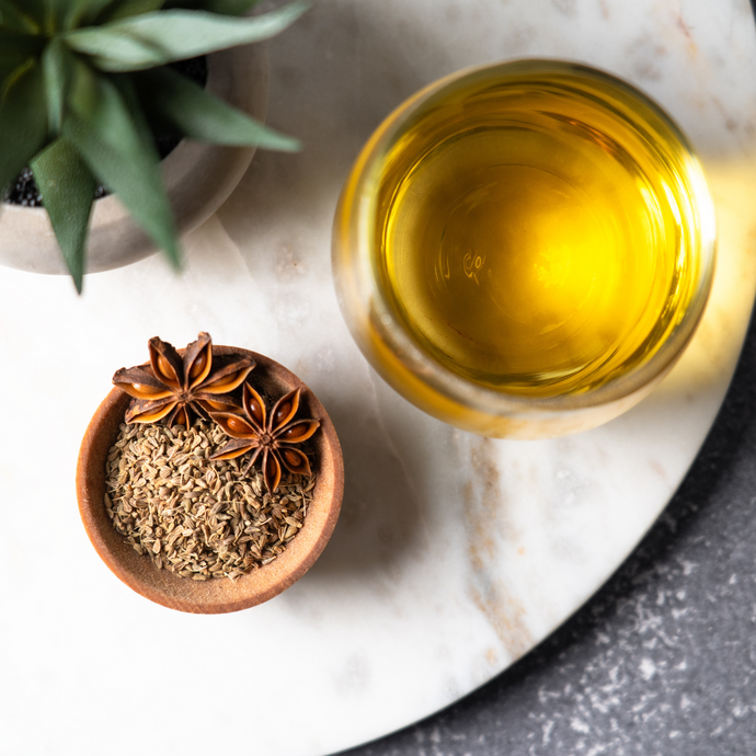 Overhead view of a glass cup of Organic Anise Seed Tea with a rich golden hue, next to a small wooden bowl filled with dried anise seeds and star anise pods on a marble board.