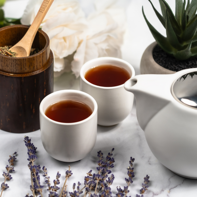 Two cups of Organic Relieve Stress Tea served with a white teapot, surrounded by lavender sprigs, flowers, and loose leaf tea in a wooden jar on a marble surface.