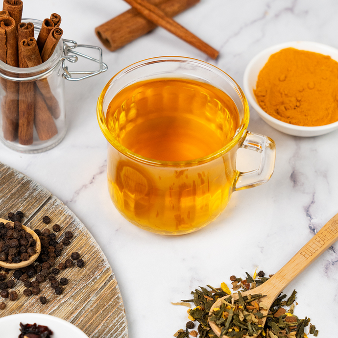 Cup of golden anti-inflammatory tea surrounded by cinnamon sticks, turmeric powder, black pepper, and loose leaf tea blend on a marble surface.