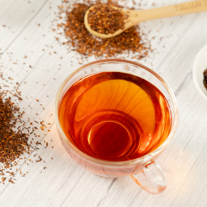 Cup of freshly brewed Organic Rooibos Tea with a warm amber hue, surrounded by scattered rooibos leaves and a wooden tea scoop on a light wooden surface.