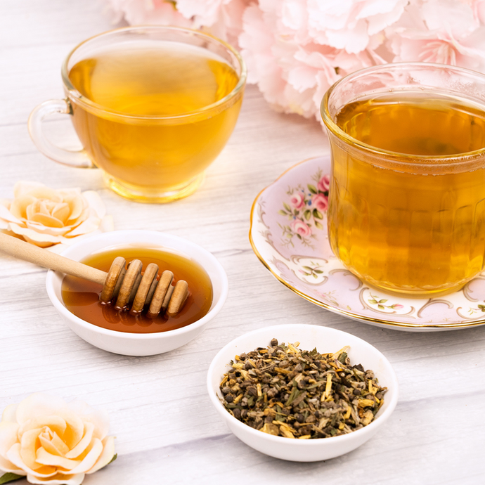 Two cups of brewed Organic Menopause Tea with a dish of honey, a bowl of loose herbal blend, and delicate flowers arranged on a light wooden surface for a calming, feminine presentation.