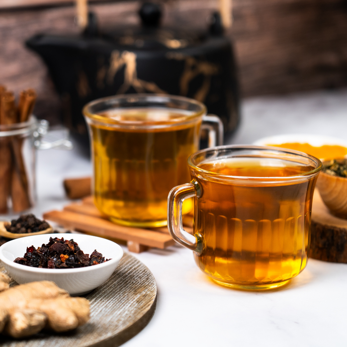 Two glass cups of Organic Anti-Inflammatory Tea with a teapot in the background, surrounded by ginger, cinnamon sticks, and dried herbs on a marble surface.