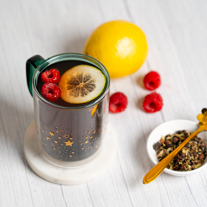Glass mug with hot beverage, lemon, and berries on a white surface with a bowl of tea leaves and honeycomb.