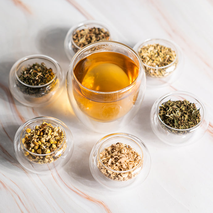 Glass cup of Organic Bloat Relief Tea surrounded by small bowls of loose herbal ingredients on a marble surface.