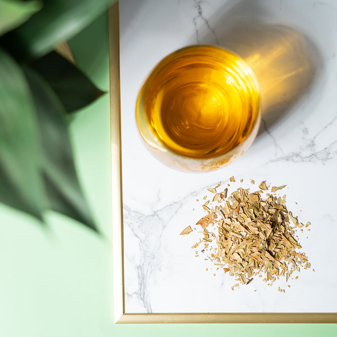 A glass cup of golden Organic Boldo Tea on a marble tray with a pile of dried boldo leaves beside it, styled with a touch of greenery.