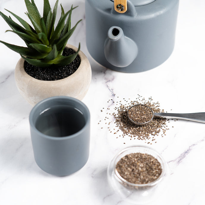 Tea set with gray teapot, cup, and small bowl of chia seeds on a marble surface.