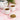 Two white cups of brewed herbal tea beside a small dish of dried chickweed leaves and a white teapot, set on a pink marble surface with greenery in the background.