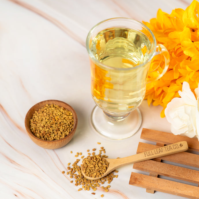 Glass cup of Organic Fenugreek tea with a wooden spoon and bowl of fenugreek seeds on a light marble surface with yellow flowers.