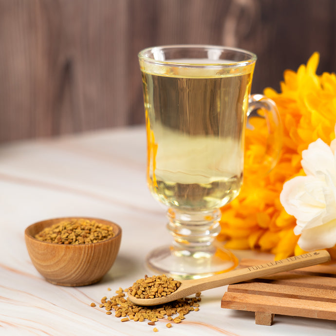 Glass of fenugreek tea with a wooden bowl and spoonful of fenugreek seeds on a wooden surface.