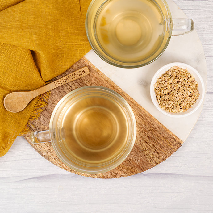 Two clear glass mugs with tea, a wooden spoon, and a small bowl of ginger root on a white surface.