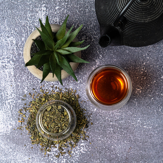 Tea-making setup with a cup of tea, dried herbs, a teapot, and a small plant on a textured surface.