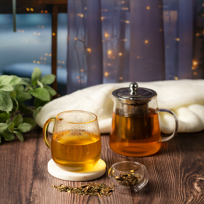 Tea set with glass mug, teapot, and tea leaves on a wooden table with a cozy background.