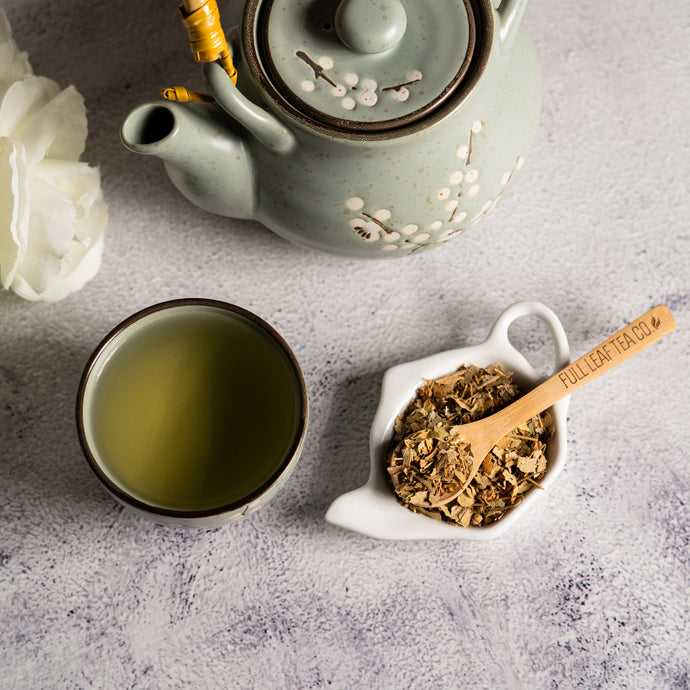 Tea set with a teapot, cup of tea, and container of linden leaf leaves on a textured surface.