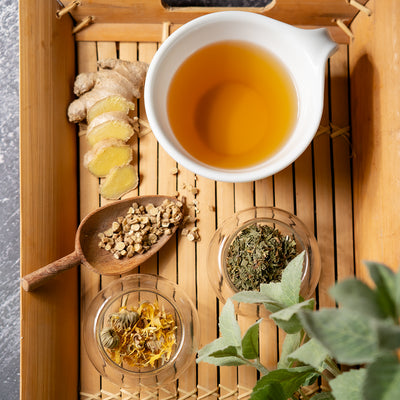 Tea-making setup with a bowl of tea, ginger slices, and herbs on a wooden tray.