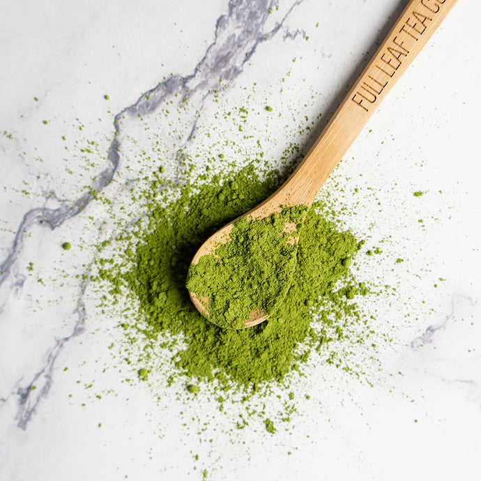 A wooden spoon engraved with “Full Leaf Tea Co.” resting on a pile of vibrant green matcha powder, with loose powder scattered across a white marble surface.