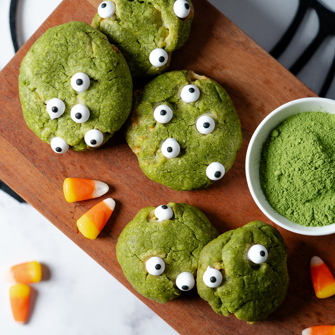 Green Halloween cookies made with Culinary Matcha, decorated with candy eyes to look like monsters, displayed on a wooden board with candy corn and a bowl of vibrant green matcha powder.