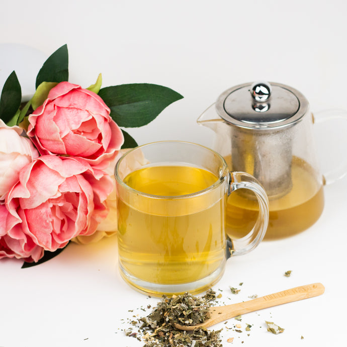 Glass mug of tea with a teapot, pink flowers, and a wooden spoon with tea leaves on a white background