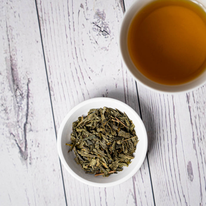 Dry sencha green tea leaves in a white bowl with a cup of brewed tea on a wooden surface