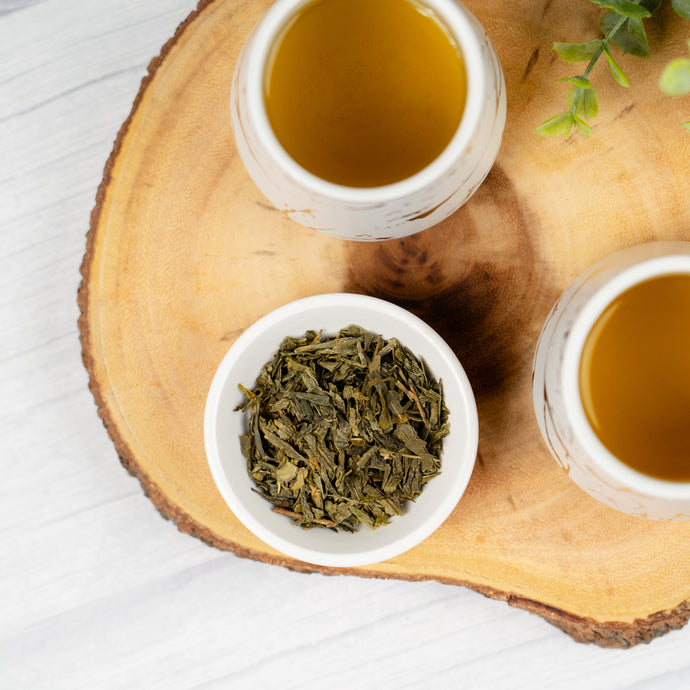 Two cups of tea and a bowl of dry sencha tea leaves on a wooden board.