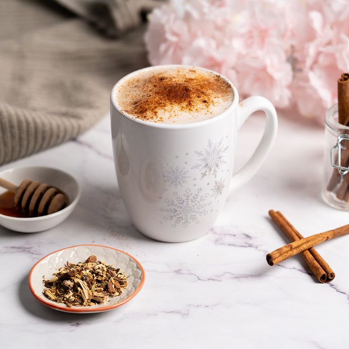 White mug with a hot beverage, cinnamon sticks, and a small bowl of honey on a marble surface.