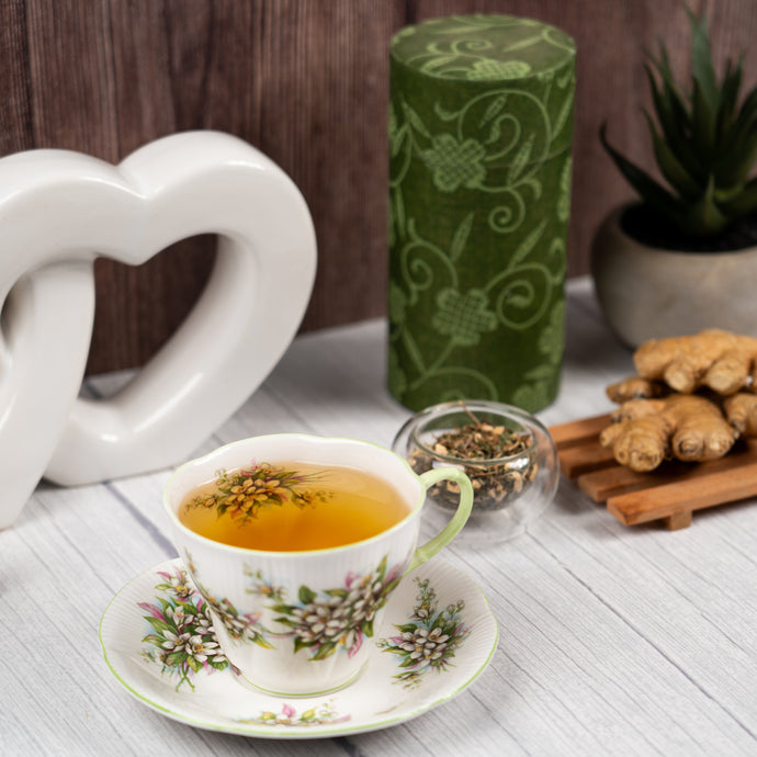 Tea cup with floral design on a white surface, surrounded by a green tea canister, ginger root, and cinnamon sticks.