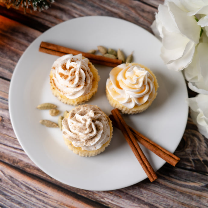 Three small cupcakes with cream cheese frosting on a white plate with cinnamon sticks and flowers on a wooden surface.