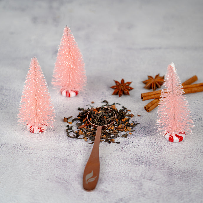 A festive flat lay with a wooden tea scoop resting on Organic Holiday Cheer loose leaf tea and spices, surrounded by cinnamon sticks, star anise, and three small pink decorative Christmas trees on peppermint bases.