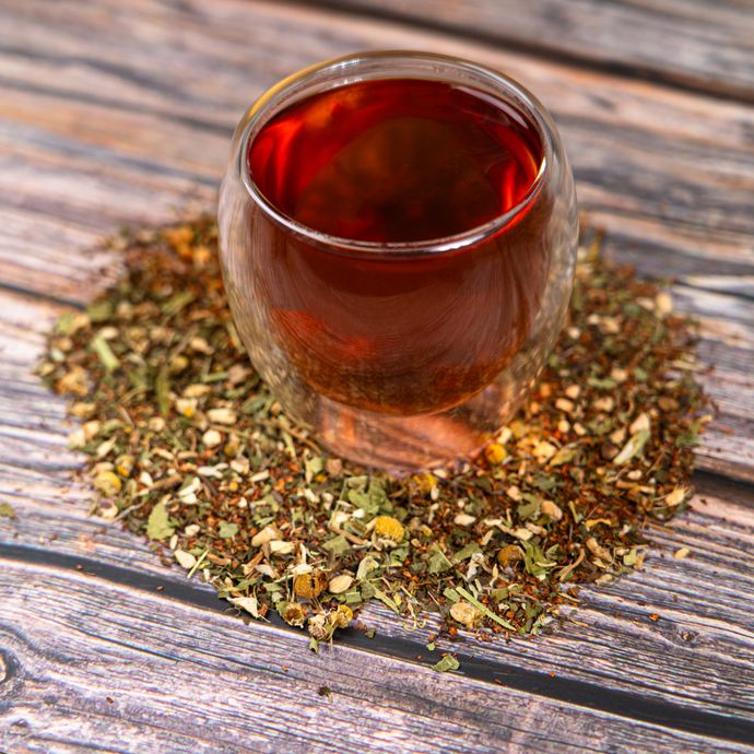 A clear double-walled glass filled with brewed Organic Fresh Start Tea, surrounded by loose herbal tea leaves and chamomile flowers on a rustic wooden surface.