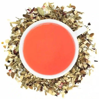 A cup of Organic Blood Pressure Tea with a bright red brew, surrounded by dried herbs and spices used in the tea blend.