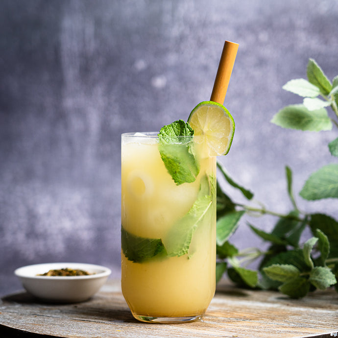 A tall glass of iced Organic Recovery Tea garnished with mint leaves and a lime slice, served with a bamboo straw, on a rustic surface with fresh mint in the background.