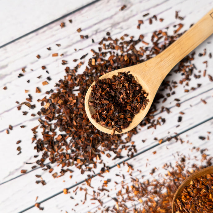 A wooden spoon filled with loose honeybush tea leaves on a white wooden surface, with more tea scattered around.