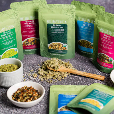 Assortment of Full Leaf Tea Co. organic teas in green pouches, including Green Energy, Vita Me, Mullein, Detox, and Pink Tea, with bowls and a wooden spoon.