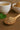Wooden spoon with tea leaves, a cup of tea, and a bowl of tea leaves on a wooden surface.