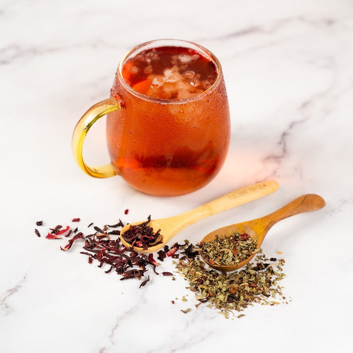 Glass mug with nw sunset iced tea, wooden spoons with tea leaves, and dried tea leaves on a marble surface.