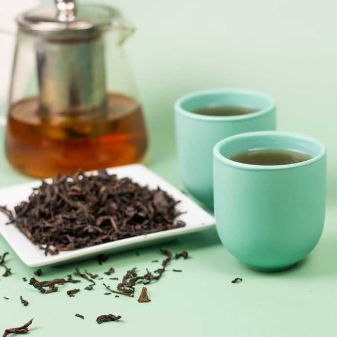 Tea set with two green cups, a teapot, and a plate of oolong tea leaves on a light green background.