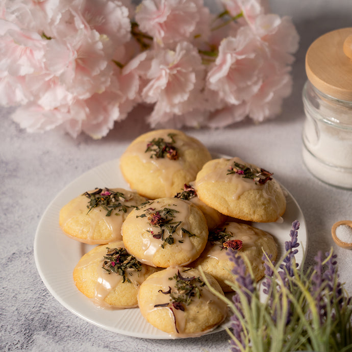 A plate of glazed cookies topped with loose leaf pink tea and flower petals, styled with pink blossoms, lavender sprigs, and a glass jar of sugar in the background.