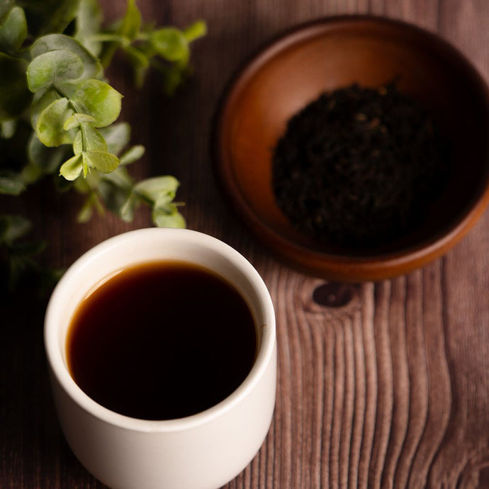 White cup of black coffee on a wooden table with a bowl of tea leaves and a plant.