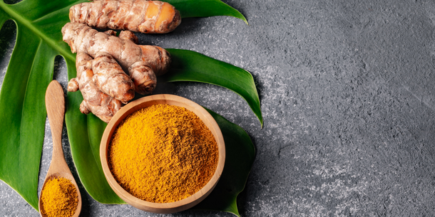 A bowl of turmeric besides turmeric roots on top of a leaf and rocky surface.
