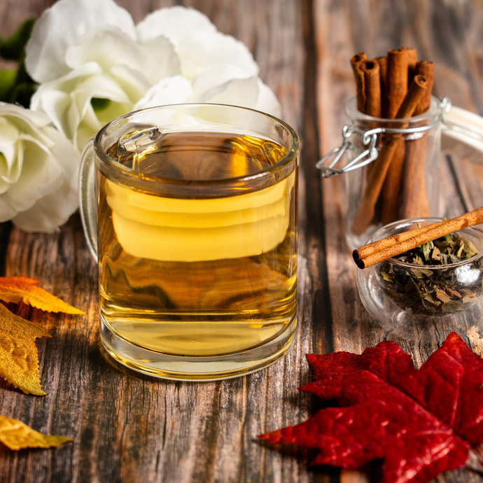 A glass mug of brewed Organic White Chai Tea on a rustic wooden table with cinnamon sticks, loose tea leaves, and autumn leaves beside white flowers.