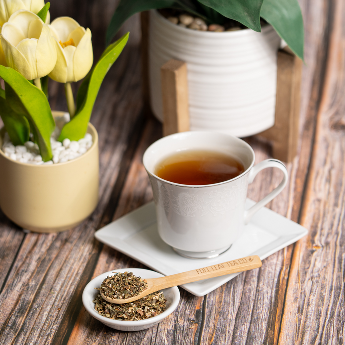 A cup of Organic Wild Lettuce Tea in a white teacup and saucer, placed beside a small bowl of loose leaf tea with a wooden Full Leaf Tea Co. spoon, surrounded by potted plants on a rustic wooden table.